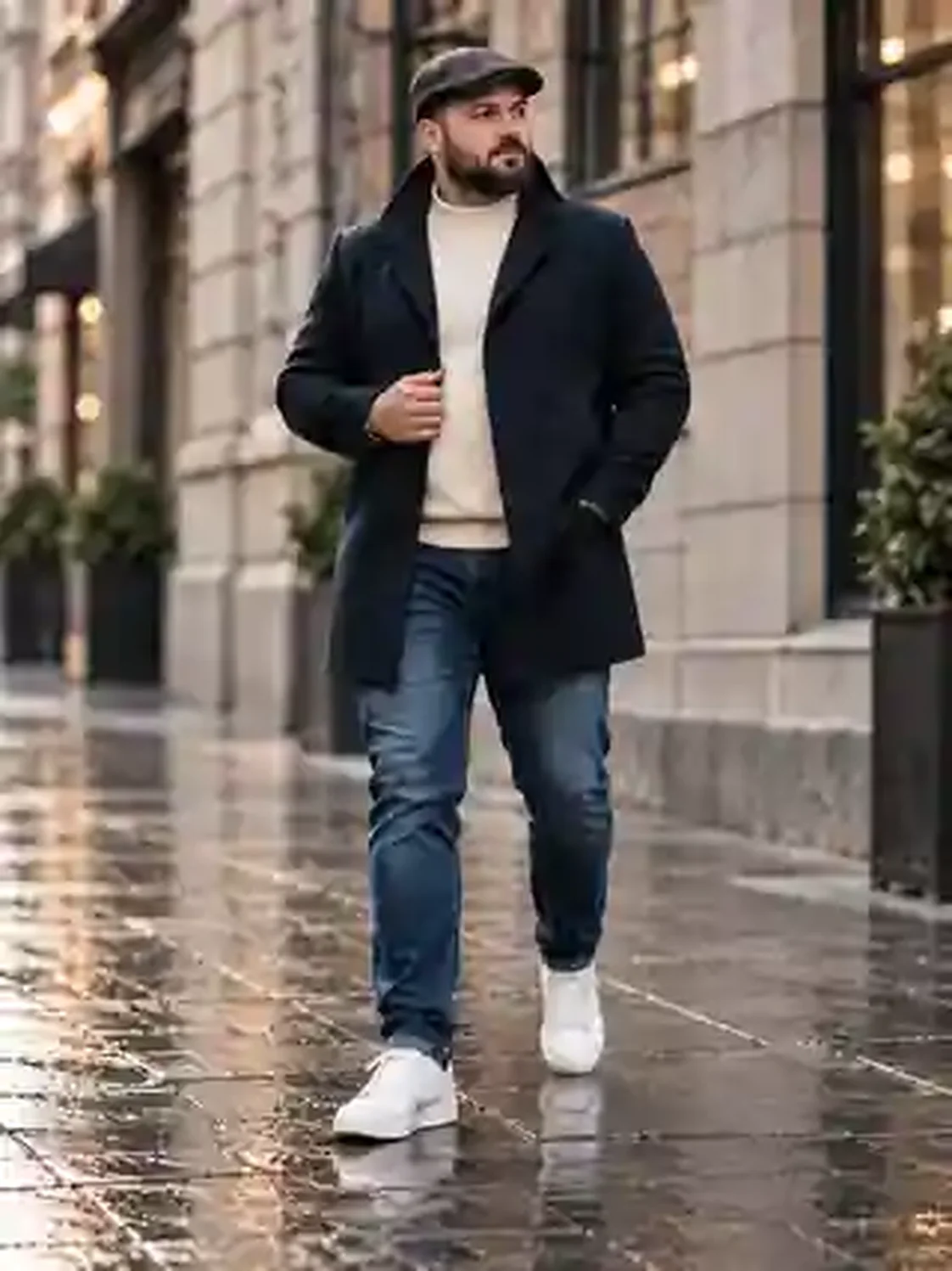 Großes bearded XXL-Model mit navyblauem Wollmantel, cremefarbenem Rollkragen, Jeans, weißen Sneakern und Schiebermütze auf nassem Bürgersteig.